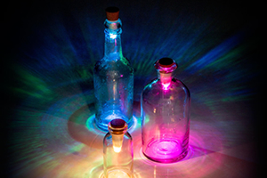 Three empty glass bottles on a table in a dark room. Each one has a bottle cork light inserted. One is glowing blue, one glowing pink and the other glowing orange.