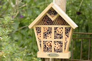 A classy house-shaped insect hotel hanging from a wooden pole. Featuring a slanted roof and several compartments.