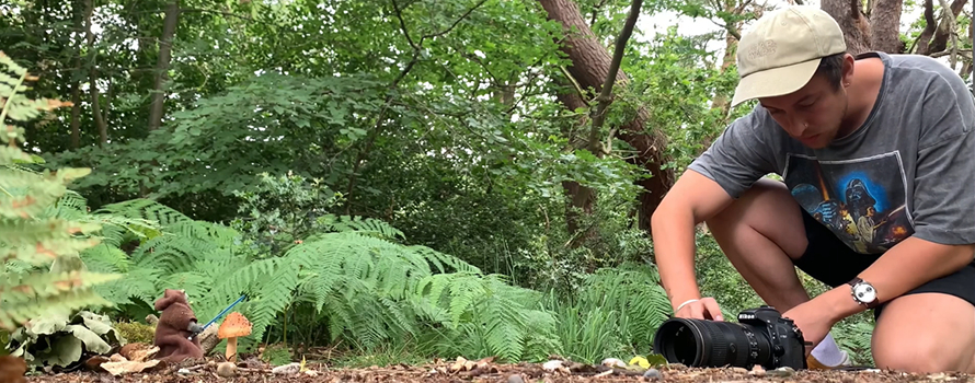 Ezra appears surrounded by woodland. He is kneeling on the ground looking at his camera, which is placed on the ground. The camera is aimed at a small Ewok Jedi figure, which is holding a blue lightsaber and standing next to a mushroom.